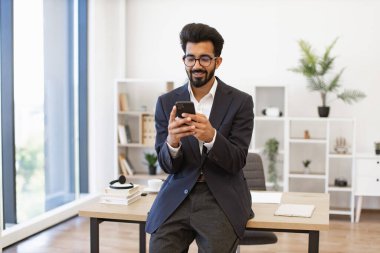 Takım elbiseli Hintli bir işadamı modern ofis ortamında akıllı telefonunu kullanırken gülümsüyor.