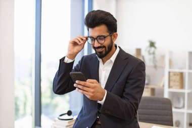 Hintli bir işadamı ofis ortamında akıllı telefonuna bakarken gülümsüyor.