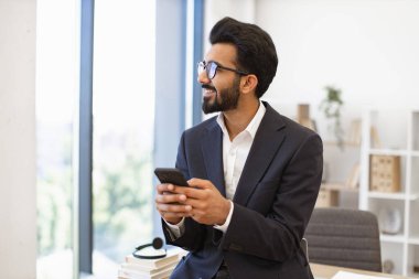 Arap bir işadamı ofis ortamında telefonunu tutarken gülümsüyor.