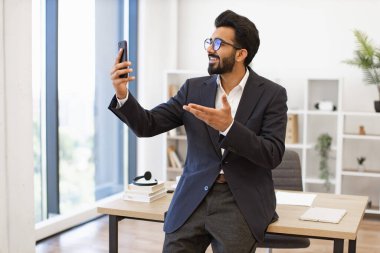 Hintli bir işadamı ofisinde bir video görüşmesi yaparken gülümsüyor, eliyle işaret ediyor.