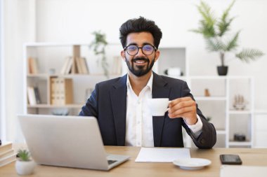 Hintli bir işadamı ofisinde bir fincan kahve tutarken gülümsüyor.