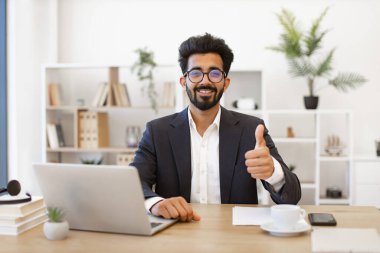 Takım elbiseli bir Hintli işadamı dizüstü bilgisayarı üzerinde çalışırken gülümsüyor ve onay veriyor.