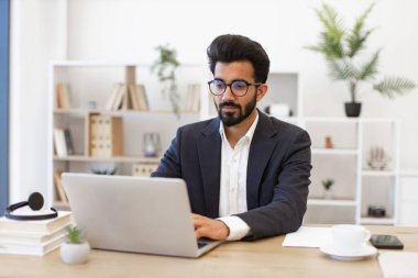 Gözlük takan Arap bir işadamı modern ofis ortamında dizüstü bilgisayar üzerinde çalışıyor.