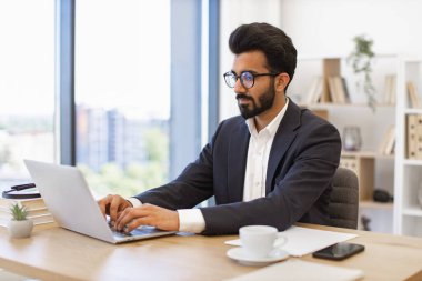 Gözlük takan Hintli bir işadamı modern ofis ortamında dizüstü bilgisayarda çalışıyor.