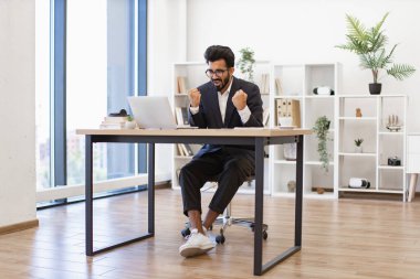 Heyecanlı bir işadamı, modern ofis ortamında dizüstü bilgisayarı üzerinde çalışırken zaferini kutluyor.