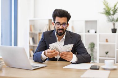 Takım elbiseli bir Hintli işadamı masasında bir tomar para sayıyor, muhtemelen başarılı bir anlaşmadan sonra.