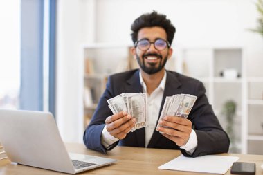 Hintli bir işadamı masasında 100 dolarlık banknotlar tutarken gülümsüyor.