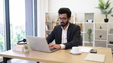 Arap bir işadamı, modern ofisinde kahve içerken dizüstü bilgisayarıyla çalışıyor.