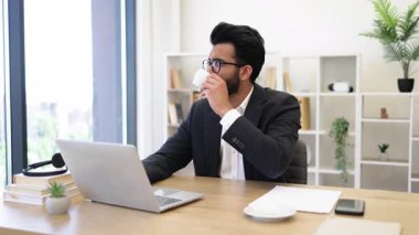 Hintli bir işadamı ofisinde bir fincan kahve içerken dizüstü bilgisayarıyla çalışıyor.