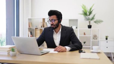 Kulaklık takan Hintli bir işadamı laptopuyla masasında çalışırken not alıyor.