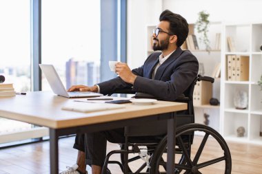 Tekerlekli sandalyedeki gülümseyen bir işadamı bir fincan kahve içerken dizüstü bilgisayarı üzerinde çalışıyor.