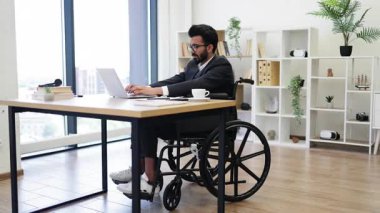 Tekerlekli sandalyedeki bir işadamı, modern ofis ortamında dizüstü bilgisayarla çalışır.