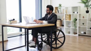 Tekerlekli sandalyedeki Hintli bir işadamı modern ofis ortamında masasında çalışıyor.