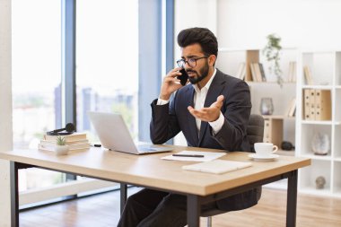 Ciddi bir Arap işadamı ofis masasında telefonuyla konuşurken el kol hareketi yapar.
