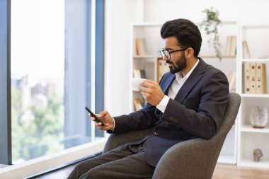 Arap bir işadamı ara veriyor, kahve içiyor ve modern bir ofis ortamında akıllı telefonunu kullanıyor.