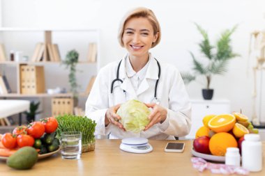 Gülümseyen bir diyetçi taze meyve ve sebzelerle çevrili bir lahana tutar, sağlıklı beslenme ve beslenmeyi teşvik eder..