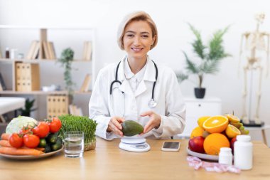 Gülümseyen bir beslenme uzmanı taze meyve ve sebzelerle çevrili bir avokado tutar ve sağlıklı beslenme alışkanlıklarını teşvik eder..