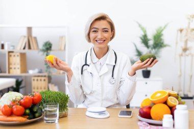 Beyaz önlüklü gülümseyen bir beslenme uzmanı taze ürünlerle çevrili bir limon ve avokado tutuyor ve sağlıklı beslenmeyi teşvik ediyor..