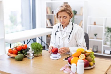 Beyaz önlüklü bir diyetçi, taze meyve ve sebzelerle çevrili kırmızı bir elmayı tartarak sağlıklı beslenme alışkanlıklarını teşvik eder..