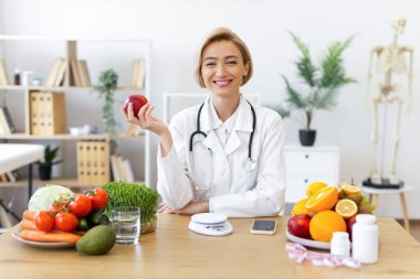 Beyaz önlüklü gülümseyen bir beslenme uzmanı, meyve ve sebzelerle çevrili bir elma tutuyor ve sağlıklı beslenmeyi teşvik ediyor..