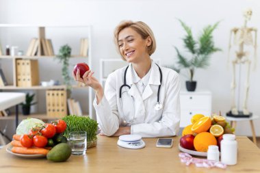 Gülümseyen bir diyetçi taze ürünlerle çevrili bir elma tutar, sağlıklı beslenmeyi ve sağlığı teşvik eder..