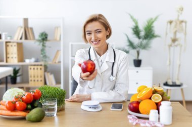 Gülümseyen bir diyetçi kırmızı bir elma sunar, sağlıklı beslenme ve beslenmeyi teşvik eder, parlak, davetkar bir ortamda.