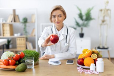 Beyaz önlüklü bir beslenme uzmanı, ahşap bir masada sağlıklı yemek seçenekleriyle çevrili kırmızı bir elmayı uzatır..