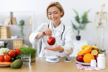 Gülümseyen bir beslenme uzmanı taze meyve ve sebzelerle çevrili kırmızı bir elma tutar ve sağlıklı beslenme alışkanlıklarını teşvik eder..