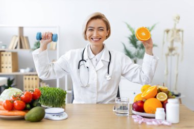 Gülümseyen bir beslenme uzmanı elinde bir dambıl ve bir portakal tutarak ofisinde sağlıklı beslenme ve egzersizleri teşvik ediyor..