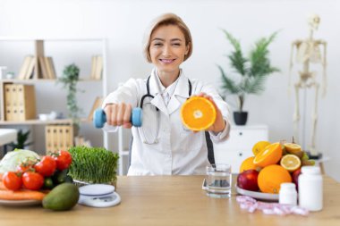 Gülümseyen bir beslenme uzmanı elinde bir portakal ve bir dambıl tutarak sağlıklı beslenmeyi ve dengeli bir yaşam tarzı için egzersiz yapmayı teşvik eder..