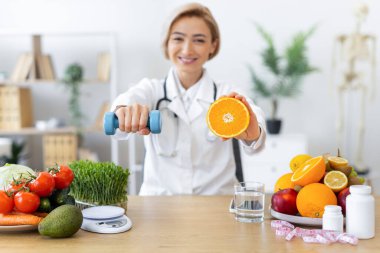 Gülümseyen bir doktor elinde bir dambıl ve dilimlenmiş bir portakal tutar. Meyve ve ve egzersizle sağlıklı bir yaşam tarzını teşvik eder..