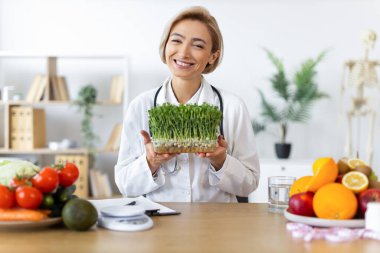 Gülümseyen bir beslenme uzmanı taze meyve ve sebzelerle çevrili ve sağlıklı beslenmeyi teşvik eden bir kap mikroyeşillik tutar..