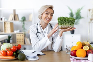 Gülümseyen bir beslenme uzmanı taze meyve ve sebzelerle çevrili, sağlıklı beslenme alışkanlıklarını teşvik eden bir kap mikroyeşillik tutar..