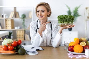 Gülümseyen bir beslenme uzmanı taze meyve ve sebzelerle çevrili bir tepsi mikroyeşile işaret ederek sağlıklı beslenmeyi teşvik eder..