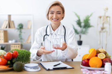 Gülümseyen bir diyetçi sağlıklı yiyeceklerle çevrili bir bardak su sunar, dengeli bir diyet ve sağlığa teşvik eder..