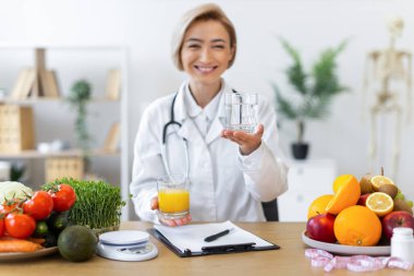 Gülümseyen bir diyetçi, taze meyve ve sebzelerle çevrili ve sağlıklı beslenme alışkanlıklarını teşvik eden bir bardak su ve meyve suyu tutar..