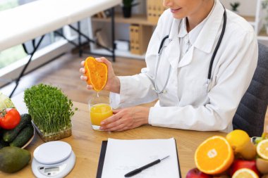 Beyaz önlüklü bir doktor sağlıklı yiyecek seçenekleriyle çevrili taze portakal suyunu bardağa sıkıyor..