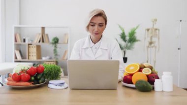Gülümseyen bir beslenme uzmanı, taze meyve, sebze ve takviyelerle çevrili bir dizüstü bilgisayarla bir masada oturur ve sağlıklı beslenmeyi teşvik eder..
