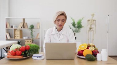 Gülümseyen bir beslenme uzmanı, elinde dizüstü bilgisayarla, taze meyve, sebze ve takviyelerle bir masada oturur..