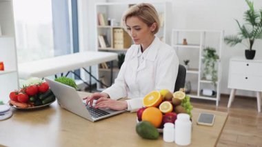 Beyaz önlüklü bir kadın, etrafında meyve, sebze ve takviyeler bulunan bir dizüstü bilgisayarda çalışır..