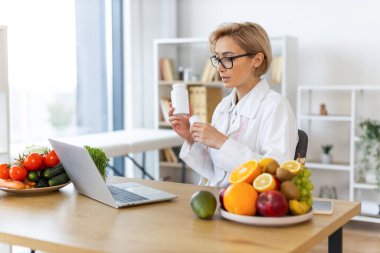 Bir beslenme uzmanı internetten bir hastaya danışmanlık yapıyor, elinde taze meyve ve sebzelerle çevrili bir ilaç şişesi tutuyor..