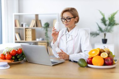 Bir kadın beslenme uzmanı, elinde destek şişeleri olan ve taze meyve ve sebzelerle çevrili bir müşterisiyle çevrimiçi olarak görüşüyor..