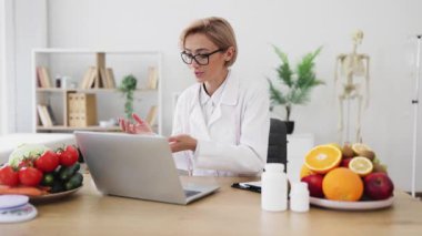 Bir beslenme uzmanı, çevrimiçi bir konsültasyon sırasında taze ürün ve dizüstü bilgisayarla çevrili sağlıklı beslenmenin faydalarını açıklar..