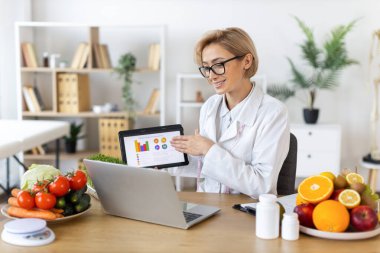 Bir dietisyen, yiyecekleri meyve ve sebzelerle çevrili bir tablet ve dizüstü bilgisayar kullanarak açıklar..