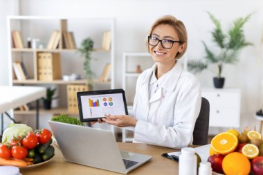 Diyetisyen bir gülümseme, taze ürünler ve dizüstü bilgisayarla çevrili bir tablet üzerinde besleyici bilgiler sunarken.