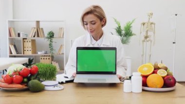 Gülümseyen bir beslenme uzmanı yeşil ekranlı bir dizüstü bilgisayar tutuyor, meyve ve sebzelerle çevrili, sağlıklı beslenmeyi teşvik ediyor..