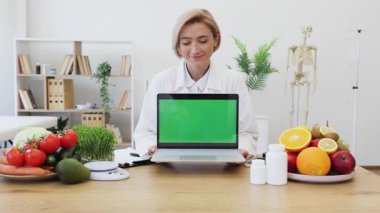 Gülümseyen bir beslenme uzmanı taze meyve ve sebzelerle çevrili yeşil ekranlı bir dizüstü bilgisayar tutuyor ve sağlıklı beslenmeyi teşvik ediyor..