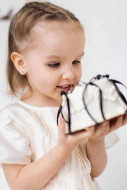 Genç bir kız elinde dekoratif bir hediyeyle gülümsüyor. Beyaz bir elbise giyiyor ve kalp şeklinde küpeleri var..