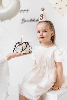 Genç bir kız üçüncü doğum gününü kutluyor, elinde mumla bir pasta tutuyor, parti şapkası takıyor ve beyaz bir yüzeye oturuyor..