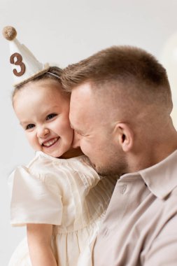 Bir baba üç numaralı doğum günü şapkasını takan gülümseyen kızını kucaklar..